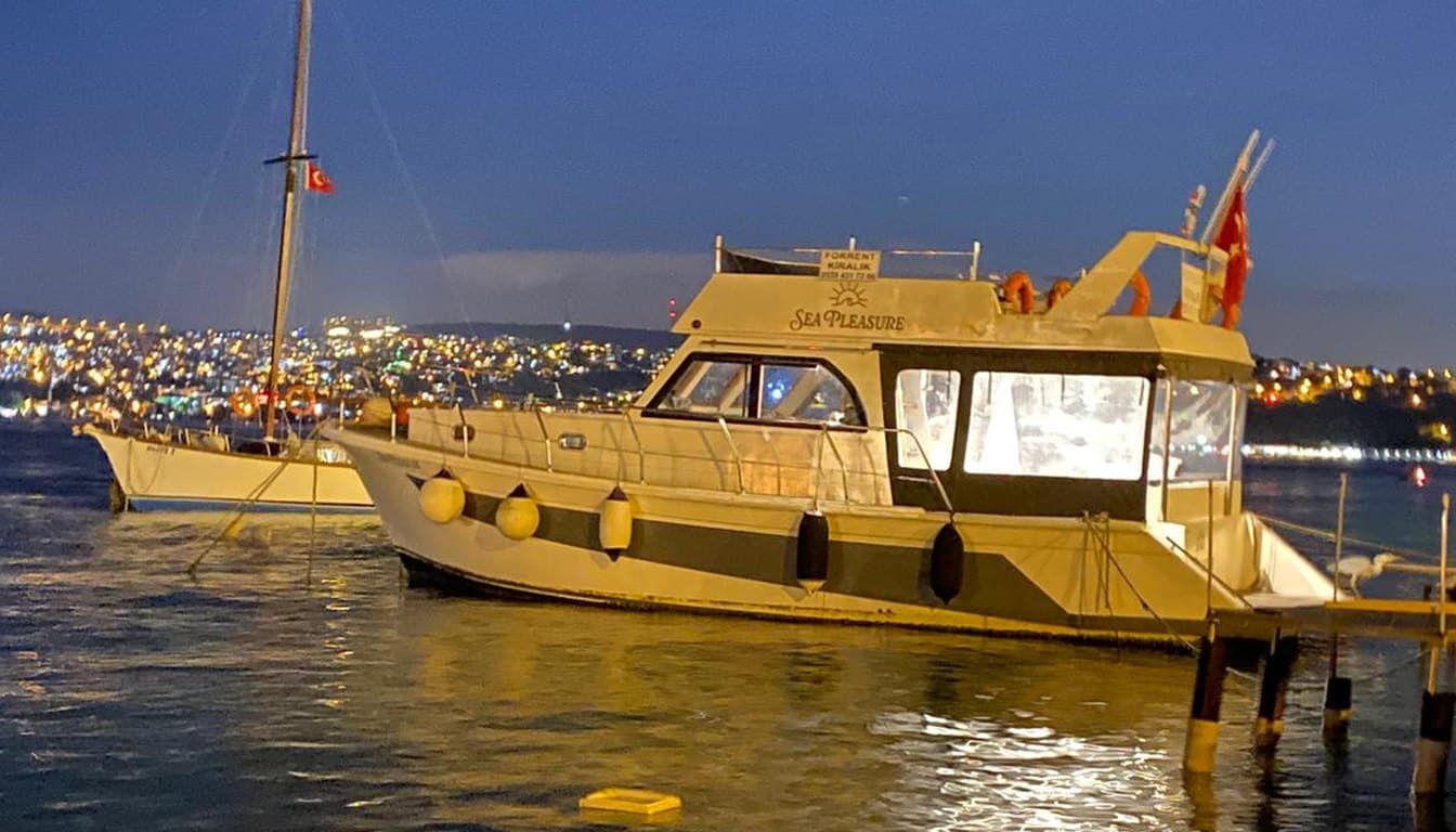 Gece İstanbul Boğazı'nda ilerleyen küçük butik özel yatın sıcak ışıklı güvertesi ve arkada aydınlatılmış köprü ile yalıların suya vuran yansımaları.